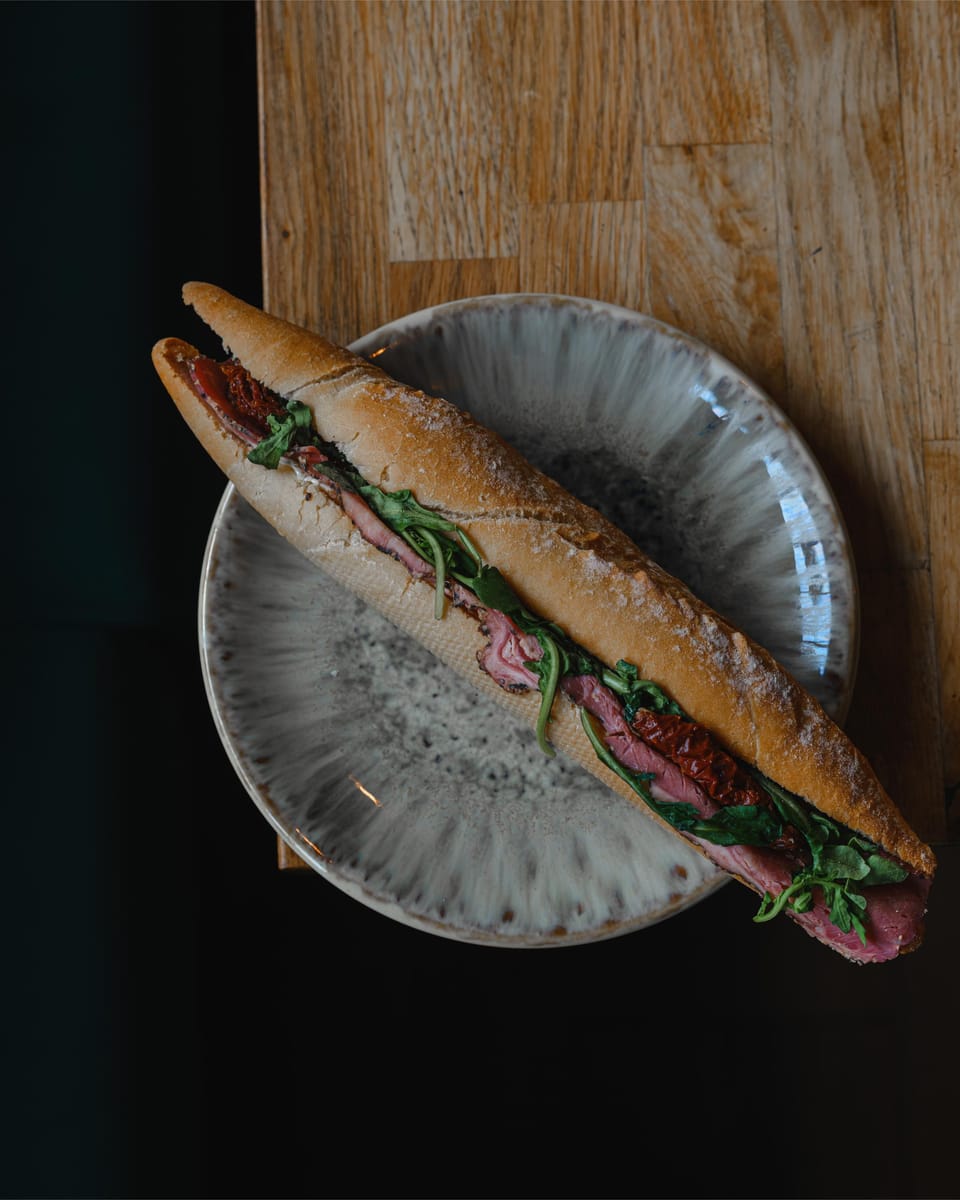 A baguette filled with sliced meat, sun-dried tomato and rocket on a grey stoneware plate.