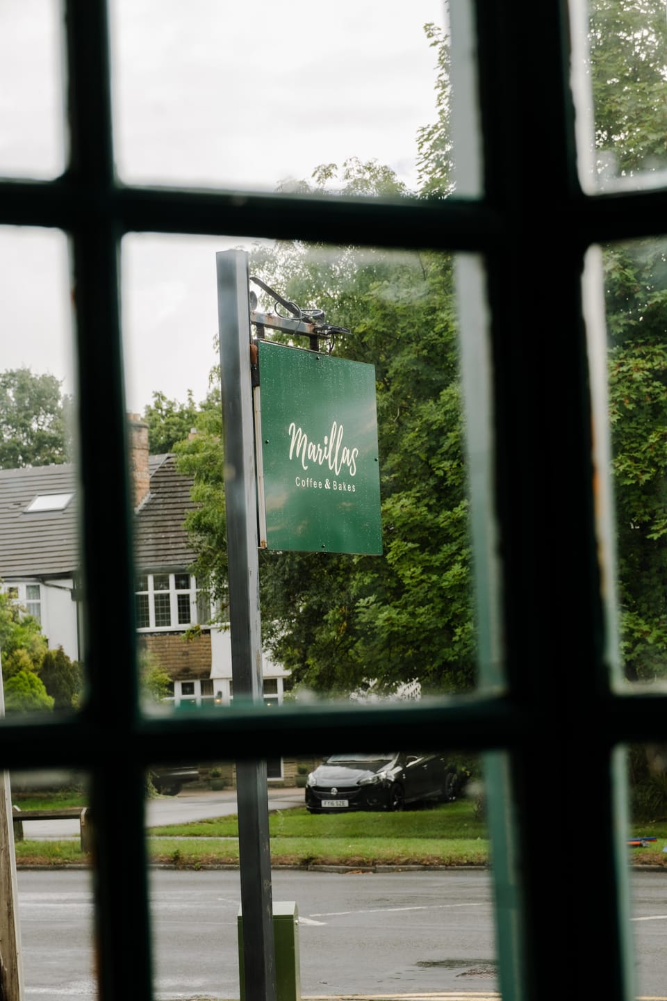 The green Marillas sign hanging outside the café, seen through a leaded window.