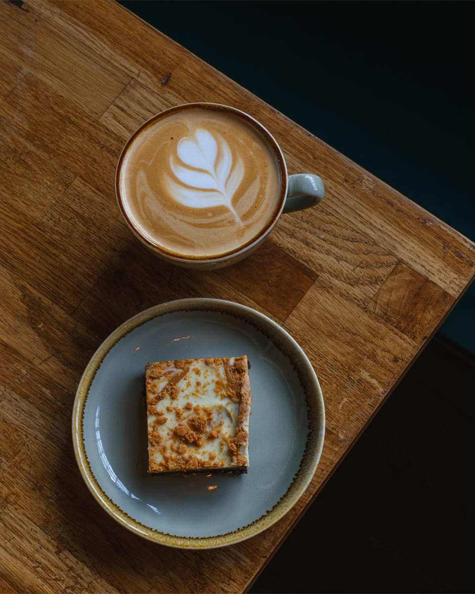 A latte with a leaf poured into the foam, beside a Biscoff brownie on a sage plate.