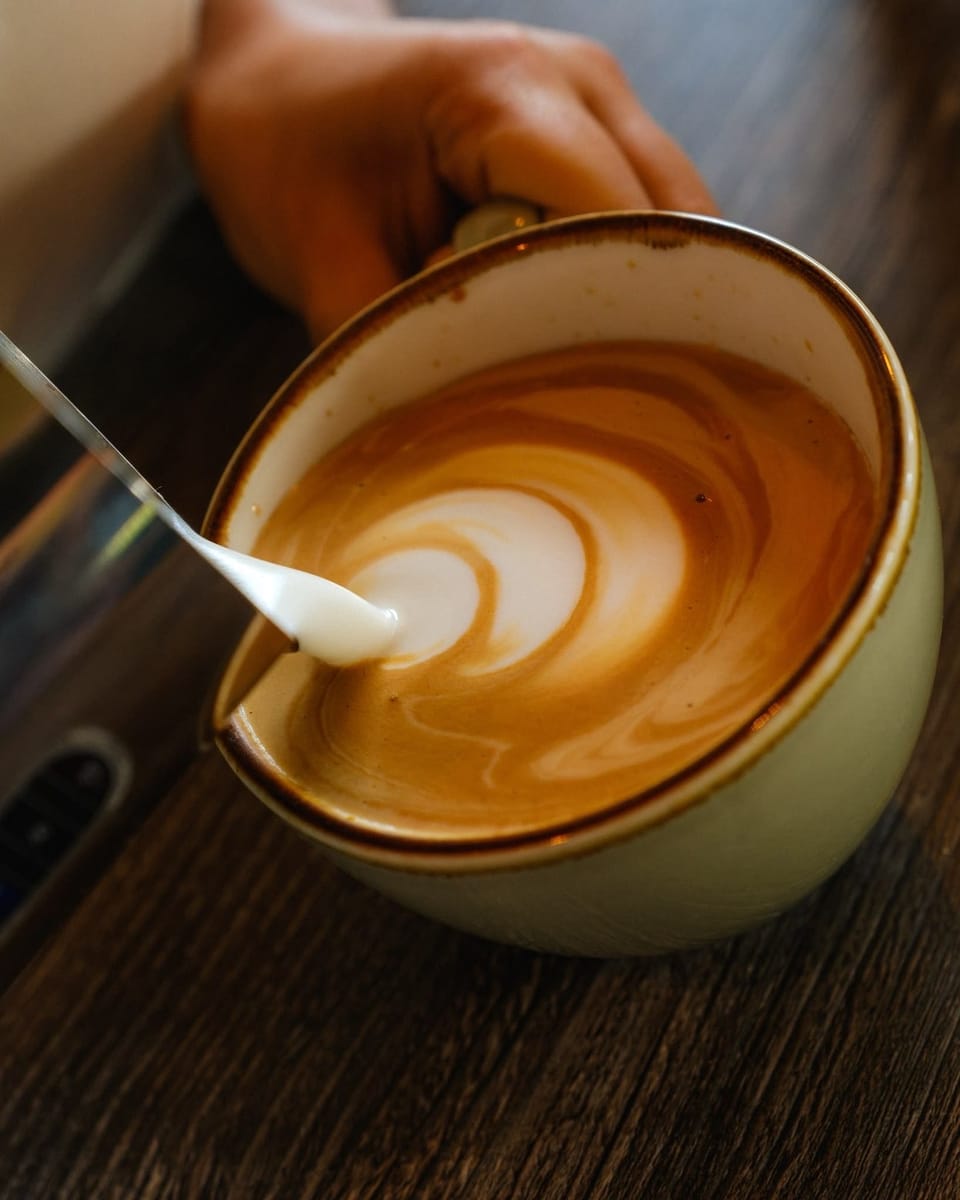 A hand holding a sage-green cup while a steel spout draws a swirl of white milk foam across a latte.