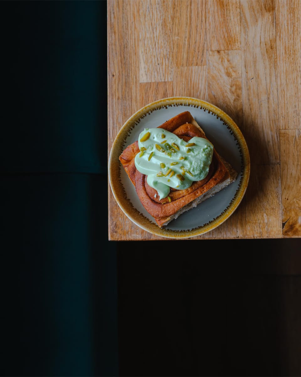 A square cinnamon bun under a thick layer of pistachio icing and chopped pistachios.