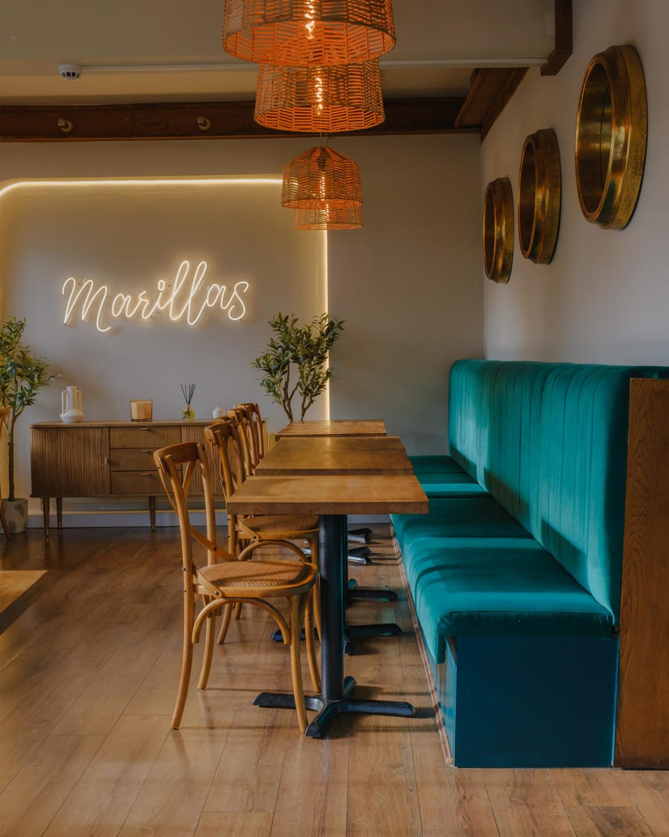 Inside Marillas: the Marillas neon sign glowing on a cream wall, rattan lamps over oak tables and bentwood chairs, and a long teal velvet banquette.