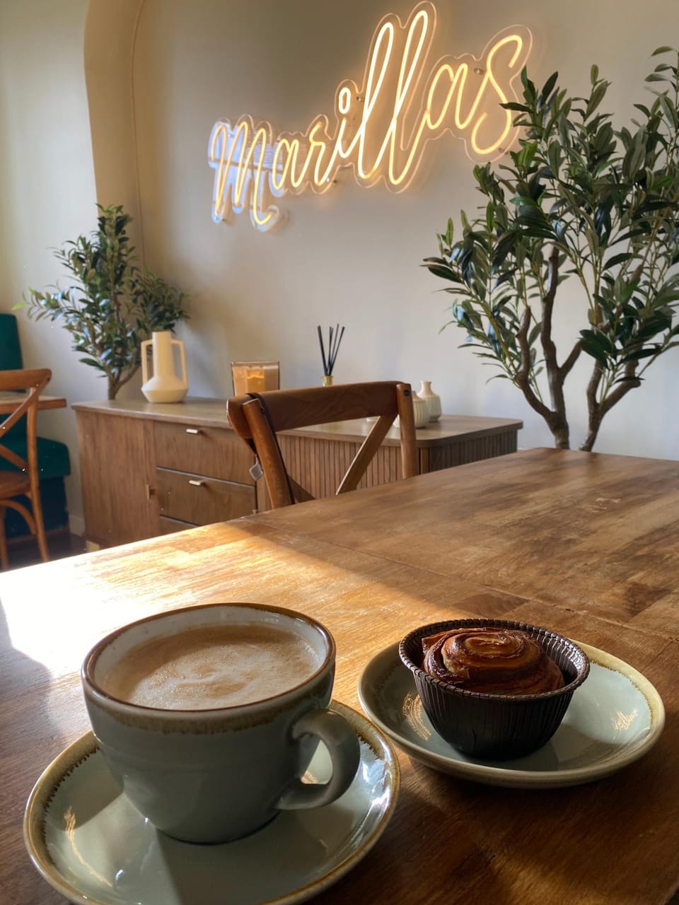 A flat white and a bake on an oak table beneath the glowing Marillas neon sign, with olive trees and an oak sideboard behind.