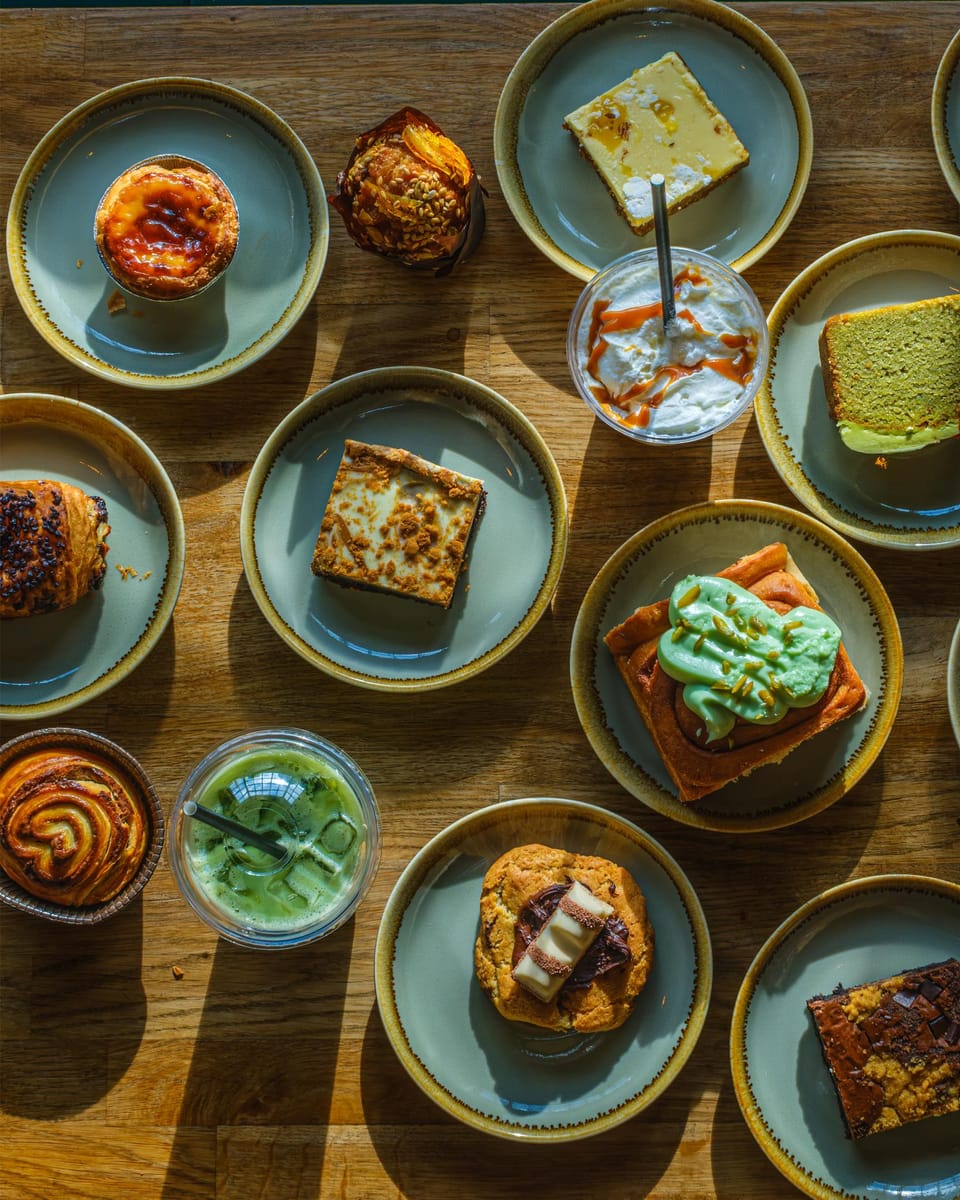 Overhead view of an oak table laid with bakes on sage plates: a pastel de nata, a muffin, a Biscoff brownie, matcha cake, a pistachio cinnamon bun, a Kinder Bueno cookie, rocky road and two iced drinks.
