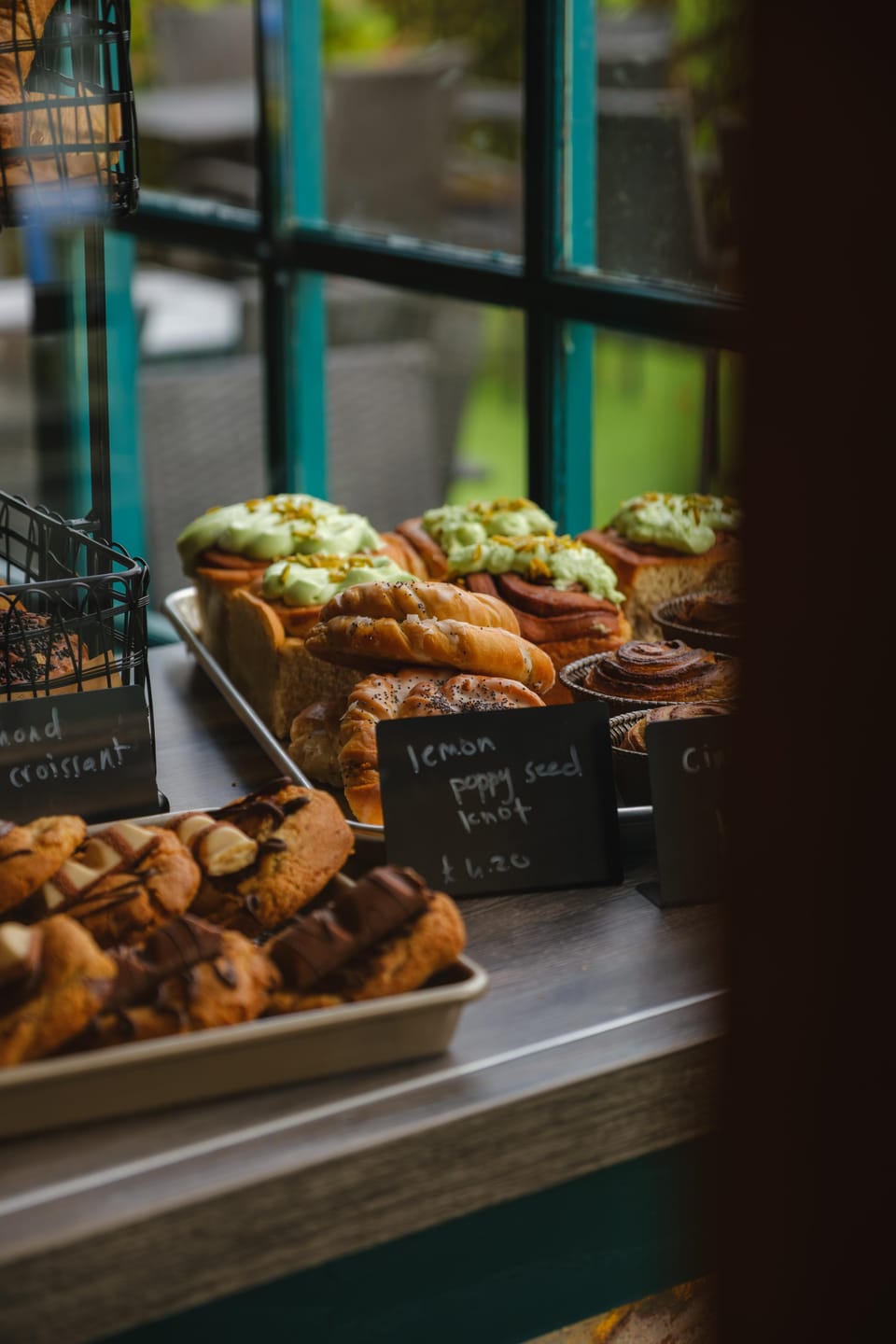 Bakes in the café window: cinnamon buns under pistachio icing, glazed pastries and a chalkboard for a lemon poppy seed knot, with green window frames behind.